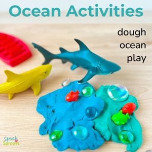 A desktop preschool ocean activity with blue dough, plastic sharks and fish, a boat and galss "stones" in the dough.